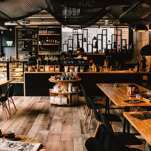A modern coffee shop interior with wooden tables, black chairs, a counter, and customers.