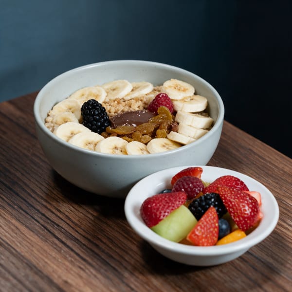 Bowl of breakfast cereal topped with bananas, berries, raisins, and a dark spread, with a side fruit bowl.