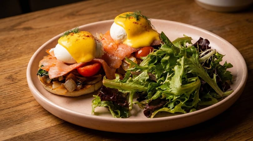 Eggs Benedict with smoked salmon, poached eggs, hollandaise sauce, and a side salad on a pink plate.