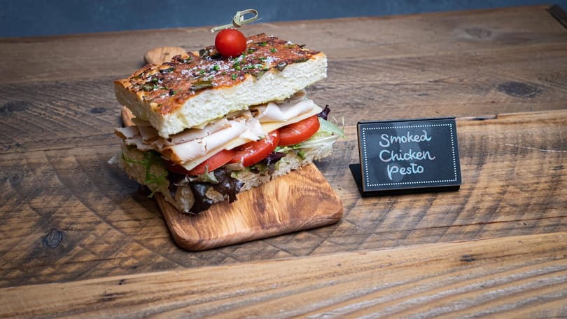 Smoked Chicken Pesto sandwich on focaccia bread with tomato, lettuce, and cheese, on a wooden board.