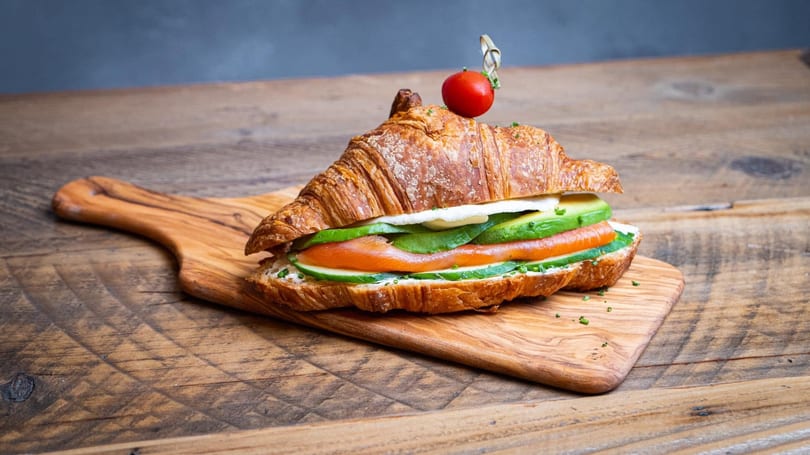 Croissant sandwich filled with smoked salmon, avocado, cucumber, and cream cheese, garnished with a cherry tomato.