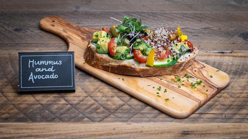 Hummus and avocado toast with tomatoes, quinoa, and microgreens on a wooden board.