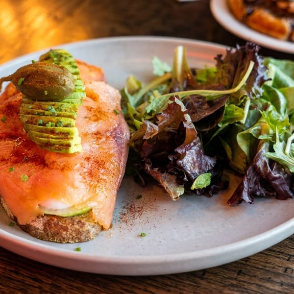 Smoked salmon toast with avocado, capers, and a side salad on a white plate.