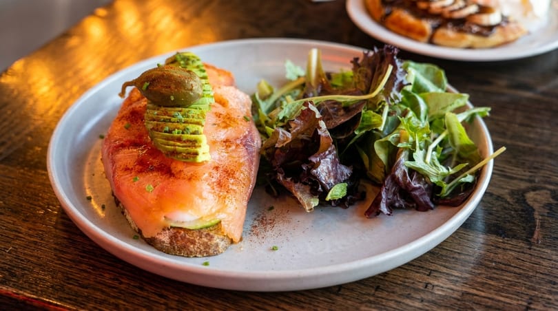 Smoked salmon toast with avocado, capers, and a side salad on a white plate.