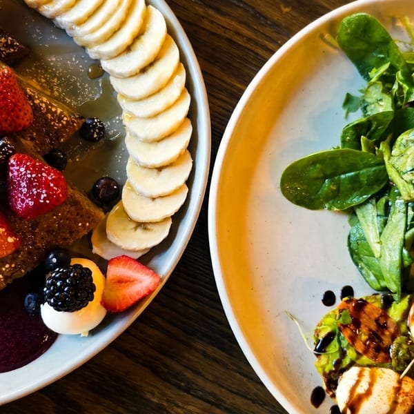 Two plates of food: one with fruit-topped pancakes and bananas, the other with a salad and caprese.
