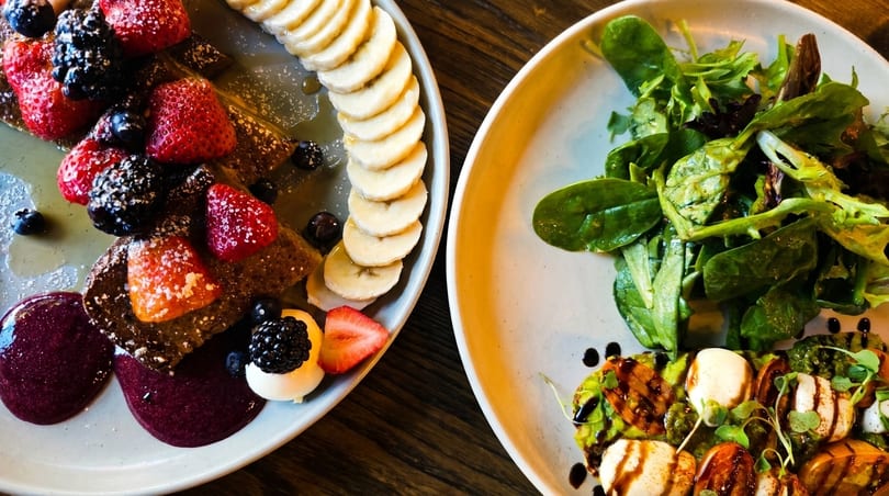 Two plates of food: one with fruit-topped pancakes and bananas, the other with a salad and caprese.