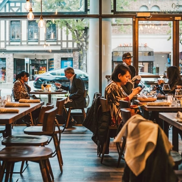 Diners enjoying meals at wooden tables in a modern restaurant with large windows overlooking a street.