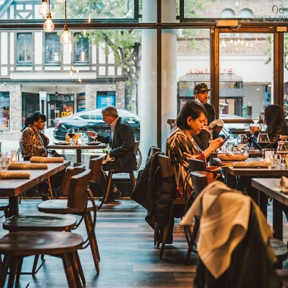 Diners enjoying meals at wooden tables in a modern restaurant with large windows overlooking a street.