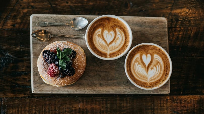 Two lattes with heart-shaped latte art and a pancake with berries on a wooden board.